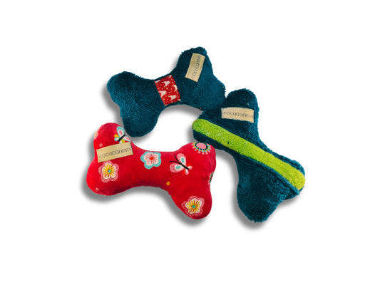 Beispielbild mit drei Plüschknochenspielzeugen für kleine Hunde. Es zeigt einen pinken Knochen mit Blumen und Schmetterlingen, einen Petrolfarbenen mit roter Borte und einen Petrolfarbenen mit einem hellgrünen Band.